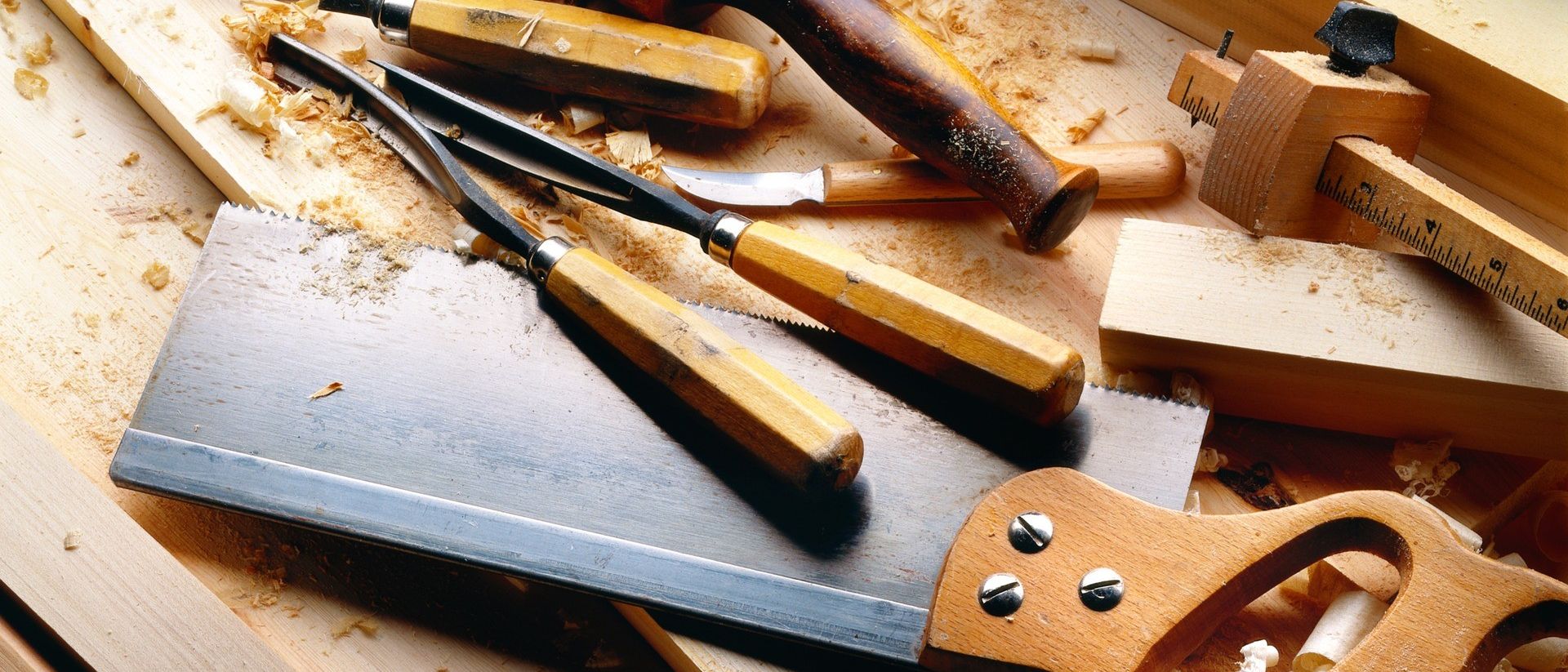 Verschiedene Holz- und Metallzimmermannswerkzeuge liegen auf einer hölzernen Oberfläche mit Sägespänen verstreut. Eine Handsäge, ein Meißel und eine Hobel sind sichtbar.