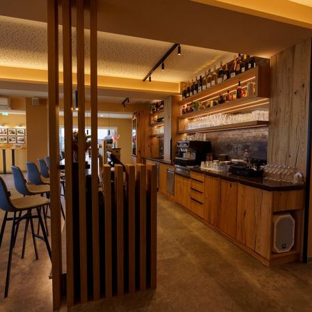 Ein Restaurantbereich mit Holzmöbeln, blauen Stühlen, einer Theke mit verschiedenen Flaschen und Weingläsern, einer Kaffeemaschine und einer hölzernen Decke mit Beleuchtung.