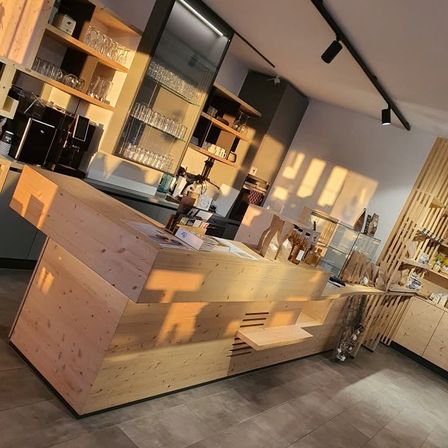 Eine Holztheke in einem Café mit Regalen und Glasvitrinen dahinter. Die Theke wird von Sonnenlicht beleuchtet.