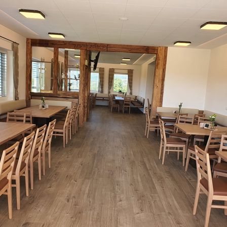 Ein leerer Essbereich mit Holztischen, Stühlen und Bänken. Der Raum hat Holzsäulen und eine Decke mit Beleuchtung. Es gibt Fenster mit Vorhängen.