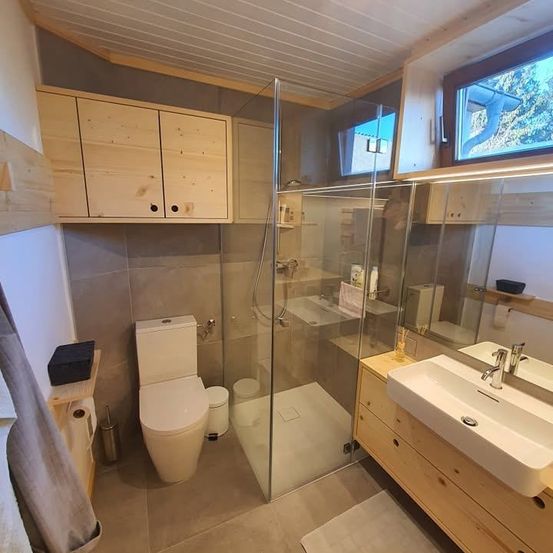 Ein modernes Badezimmer mit einer Glasdusche, einem weißen WC, einem Waschbecken und Holzschränken. Der Raum hat natürliches Licht durch die Fenster.
