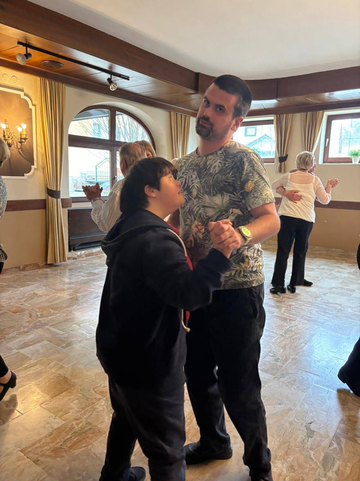 Ein Mann und eine Frau tanzen in einem gefliesten Raum mit einer Gruppe von Menschen. Der Mann trägt ein Blumenhemd und die Frau eine Kapuzenjacke.