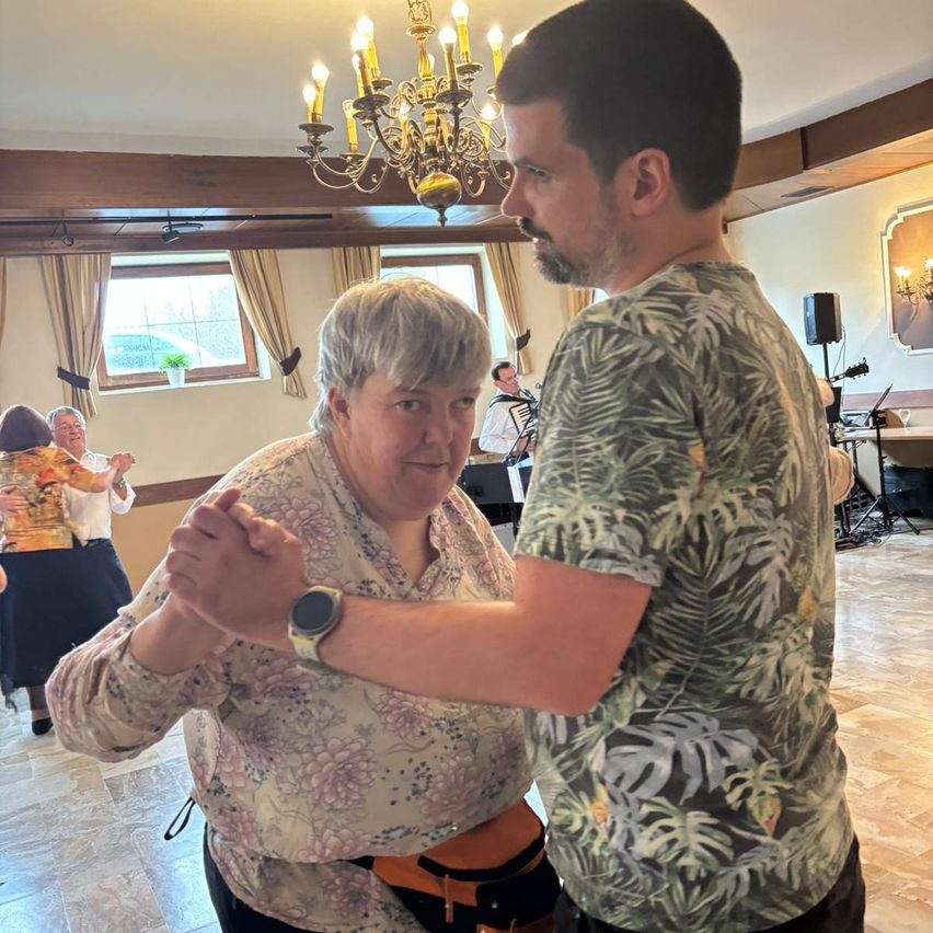 Eine ältere Frau und ein Mann tanzen zusammen in einem Raum mit einer Kronleuchter. Eine weitere Frau tanzt hinter ihnen. Ein Mann spielt Akkordeon im Hintergrund.