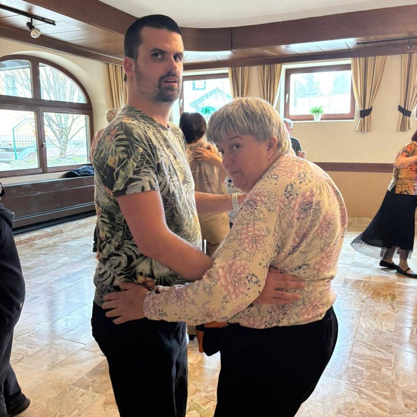 Ein Mann und eine ältere Frau tanzen in einem Raum. Sie sind von anderen Menschen umgeben, von denen einige ebenfalls tanzen.