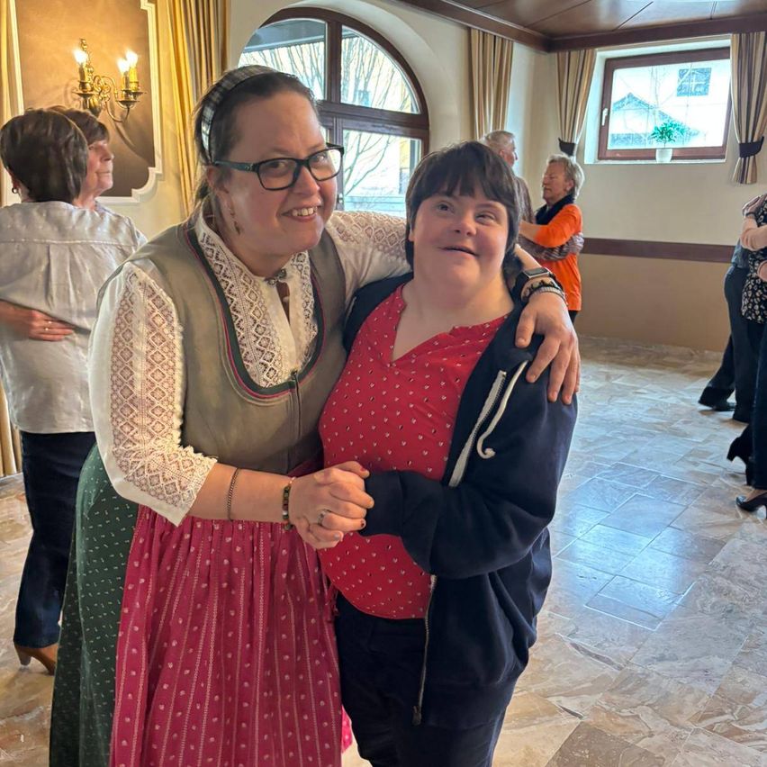 Zwei Frauen stehen eng beieinander und lächeln, eine in einem gepunkteten Kleid und die andere in einer Jacke. Hinter ihnen tanzen Menschen in einem Raum mit gefliesten Böden und einem Fenster, das Bäume draußen zeigt.