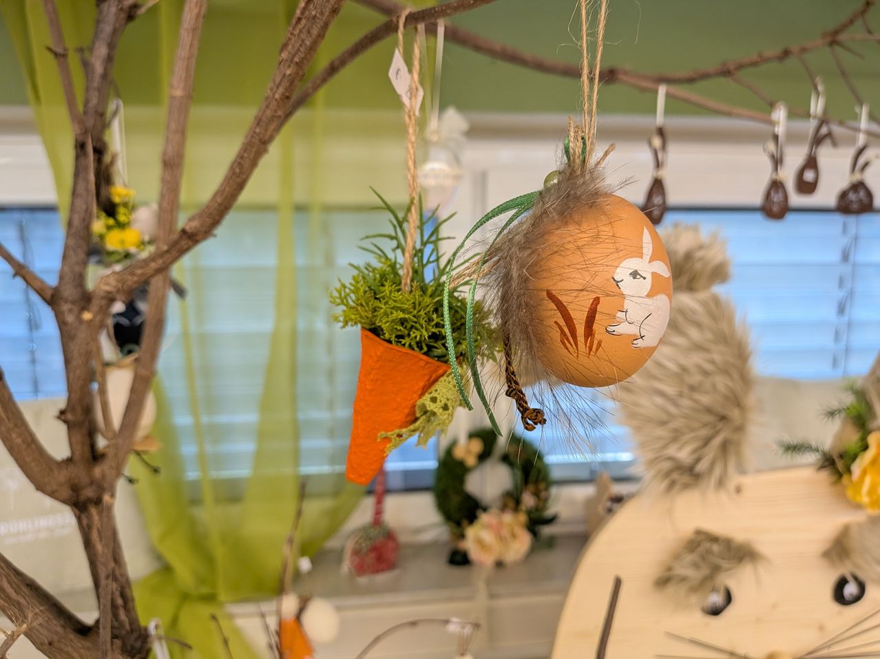 Ein Baumzweig mit Osterdekorationen hängt in einem Raum. Es zeigt ein dekoriertes Ei mit einem Hasen, einen kleinen Topf mit Pflanzen und andere Ornamente. Ein Fenster mit Jalousien und ein Holztisch mit Gegenständen sind im Hintergrund.