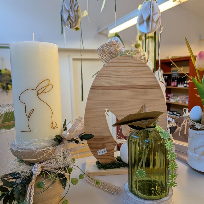 Eine weiße Kerze mit Hasendesign, ein hölzerne Ei und eine grüne Glasvase mit Blumen auf einem Tisch mit verschiedenen Dekorationen und einem Regal im Hintergrund.