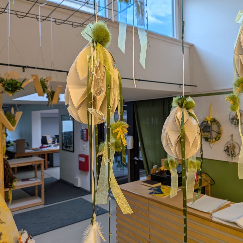 Dekorative Ostereier aus Papier hängen von der Decke in einem Raum mit einem Schreibtisch und Regalen. Der Raum hat eine weiße Decke, ein Glasfenster und eine grüne Wand.