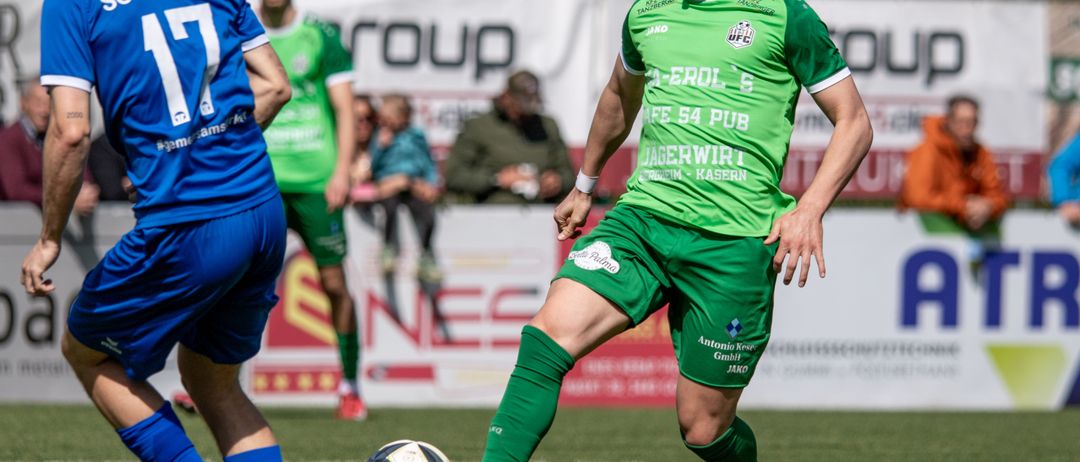 Zwei Fußballspieler in Aktion, einer in Grün und der andere in Blau. Der Spieler in Grün tritt den Ball.