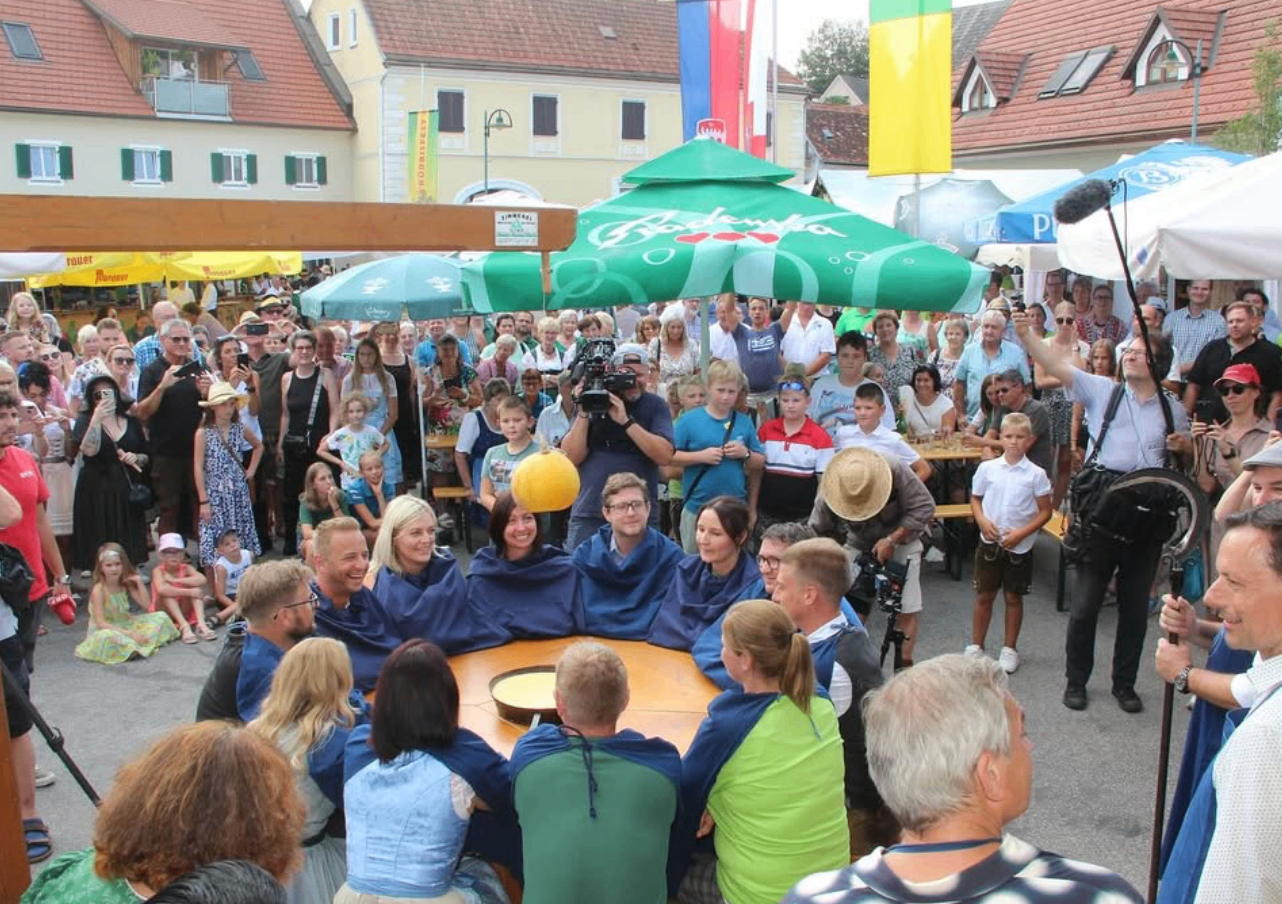 Eine Menschenmenge ist für eine Veranstaltung im Freien versammelt. Sie sitzen unter grünen Schirmen um einen Tisch herum, einige tragen blaue Umhänge. Ein Mann steht mit einem gelben Ball.