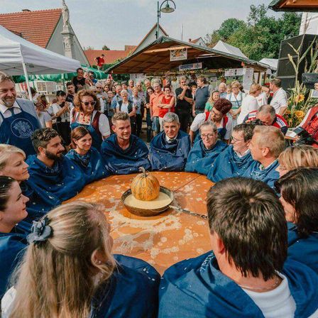 Eine Gruppe von Menschen in blauen Schürzen steht um einen Tisch mit einem Kürbis darauf. Andere Menschen stehen um den Tisch herum und einige machen Fotos.
