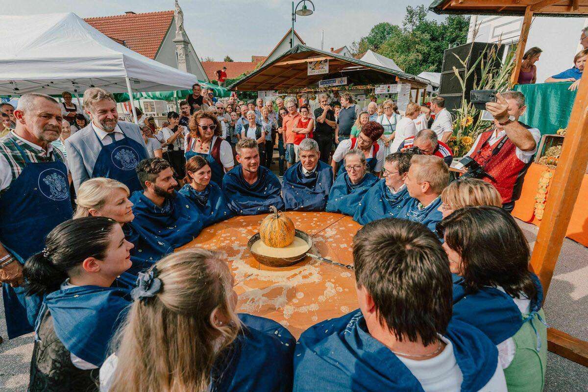 Eine Gruppe von Menschen in blauen Schürzen steht um einen Tisch mit einem Kürbis darauf. Andere Menschen stehen um den Tisch herum und einige machen Fotos.
