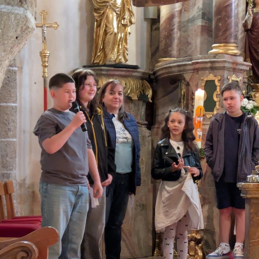 Eine Gruppe von Menschen in einer Kirche, darunter ein Junge, der in ein Mikrofon spricht, ein Mädchen, das gestikuliert, und zwei Frauen, die hinter ihnen stehen. Im Hintergrund steht eine goldene Statue.