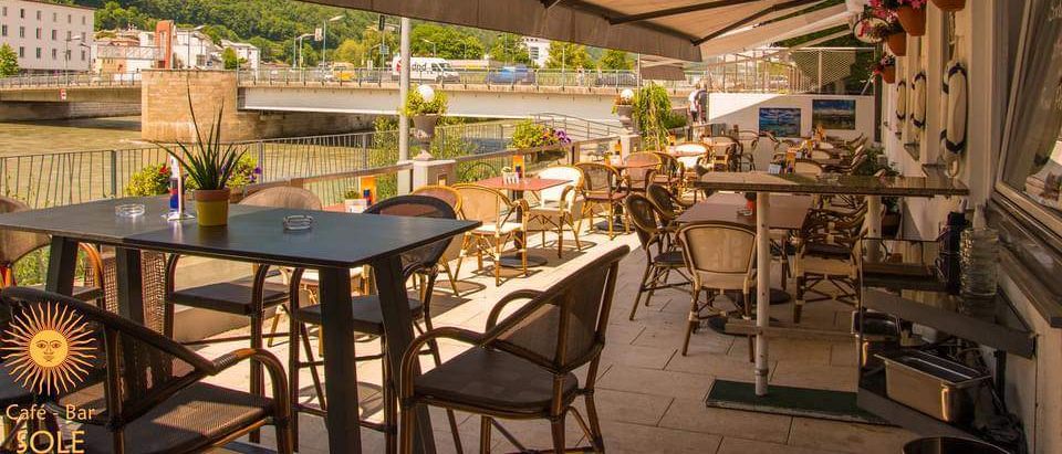 Ein Restaurant im Freien mit Tischen und Stühlen unter Markisen. Pflanzgefäße und Blumen schmücken den Bereich, und eine Brücke mit Autos und Gebäuden ist in der Ferne sichtbar.