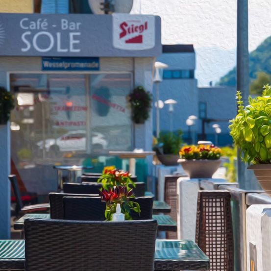 Außensitzbereich im Cafe-Bar Sole mit Tischen, Stühlen und Topfpflanzen. Ein Gebäude mit einem Schild, das Cafe-Bar Sole liest, und einem Berg im Hintergrund.