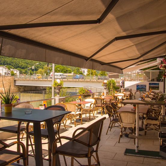 Terrasse eines Restaurants mit Tischen und Stühlen unter einem Vordach, mit Blick auf einen Fluss und eine Brücke, über die Autos fahren.