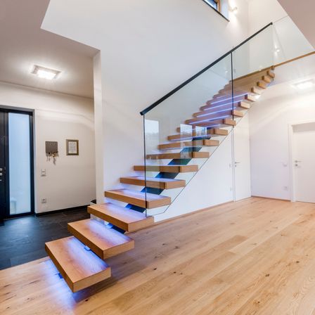 Innenansicht eines modernen Hauses mit einer Glastreppe, Holzstufen und einer weißen Wand. Blaue Lichter beleuchten die Treppe.