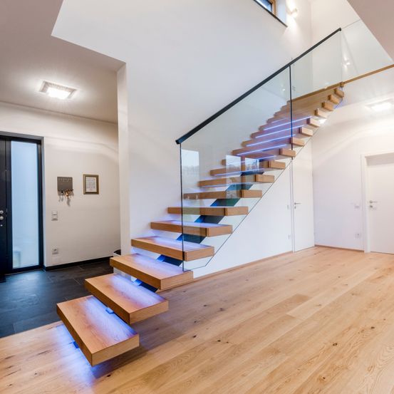 Innenansicht eines modernen Hauses mit einer Glastreppe, Holzstufen und einer weißen Wand. Blaue Lichter beleuchten die Treppe.