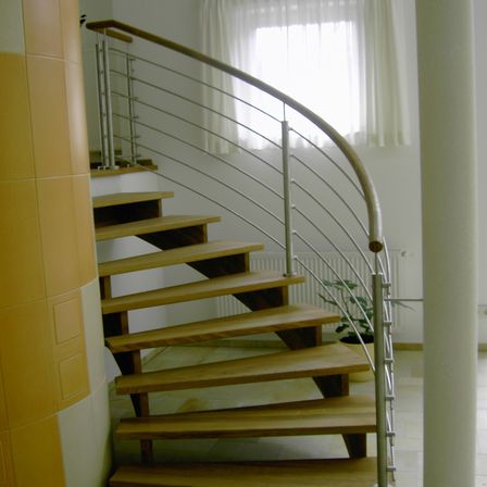 Eine Wendeltreppe mit Holzstufen und einem Metallgeländer ist zu sehen. Die Treppe wird von natürlichem Licht beleuchtet, das durch ein Fenster mit weißen Vorhängen fällt. Ein Topf mit einer Pflanze steht in der Nähe der Treppe auf dem Boden.