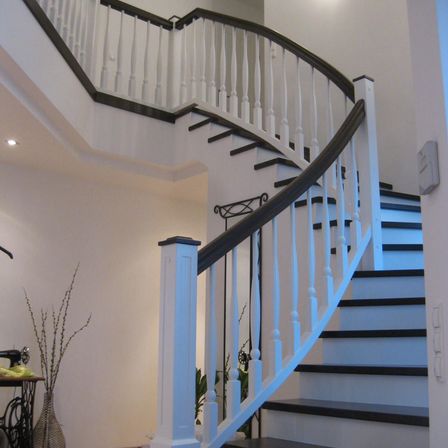 Eine Wendeltreppe mit weißen Handläufen und schwarzen Stufen. Die Wand ist weiß, und links steht eine Vase mit einer Pflanze. Eine Nähmaschine steht neben der Wand.