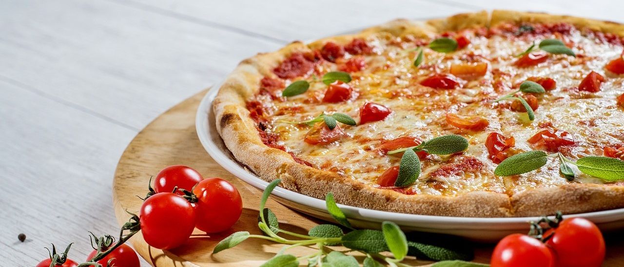 Eine runde Pizza mit geschmolzenem Käse, roten Tomaten und Salbeiblättern liegt auf einem Holzteller auf einem Holztisch.
