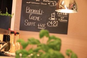 Ein schwarzes Menübrett mit 'Brokkoli Creme Suppe' für 4,40 Euro und 'Cotta' für 4,20 Euro. Darunter sind verschiedene Gewürze platziert.