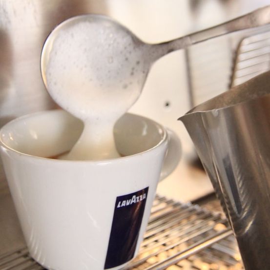 Ein Metalllöffel gießt Milch in eine weiße Lavazza-Kaffeetasse auf einem Metallrost.