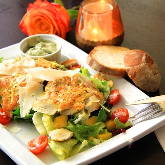 Ein Teller mit einem Salat aus gemischten Blattsalaten, Kirschtomaten und Avocado, überbacken mit Brot und gegrilltem Hähnchen. Eine Beilage Dip und eine brennende Kerze sind auf dem Tisch.