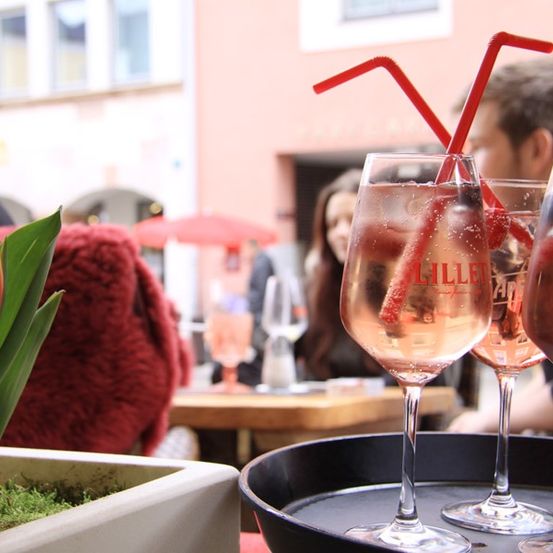 Ein Tisch mit drei Weingläsern gefüllt mit Wein, zwei mit roten Strohhalmen, einem roten Hut und einer Tulpe in einem weißen Topf.