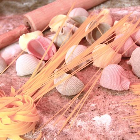 Eine Vielzahl von rosa und weißen Nudelformen liegt auf einer rosa Oberfläche verstreut. Es gibt Spaghetti, Muschelschalen und Bänder. Ein hölzerne Nudelrolle liegt links.