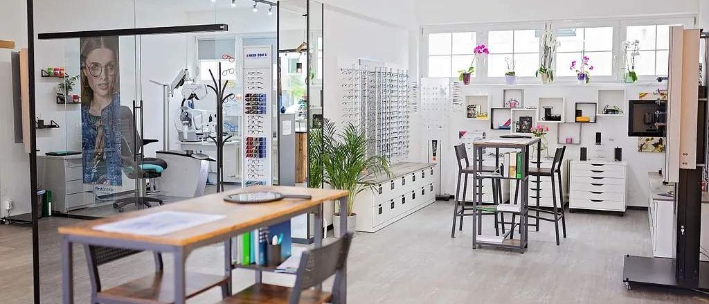 Ein gut beleuchtetes modernes Optikerbüro mit Glaswänden, Tischen, Stühlen, Schränken und Topfpflanzen. Der Raum verfügt über Regale mit gerahmten Fotos und Topfpflanzen.