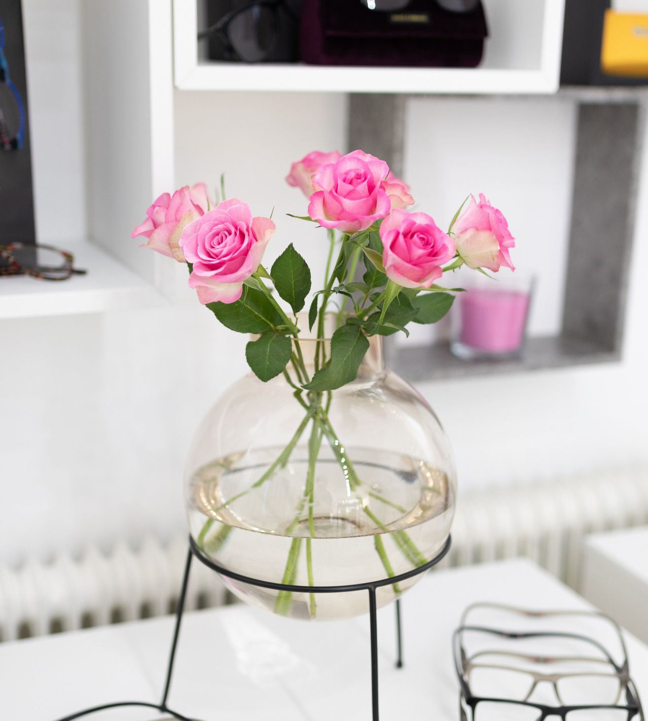 A clear glass vase filled with water and containing several pink roses sits on a white table in front of a white shelf.