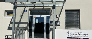 Ein Gebäudeeingang mit einer Glastür und einem Metallvordach. Ein Schild trägt die Aufschrift 'Regina P. Arztin für Allgemein' mit Kontaktinformationen.