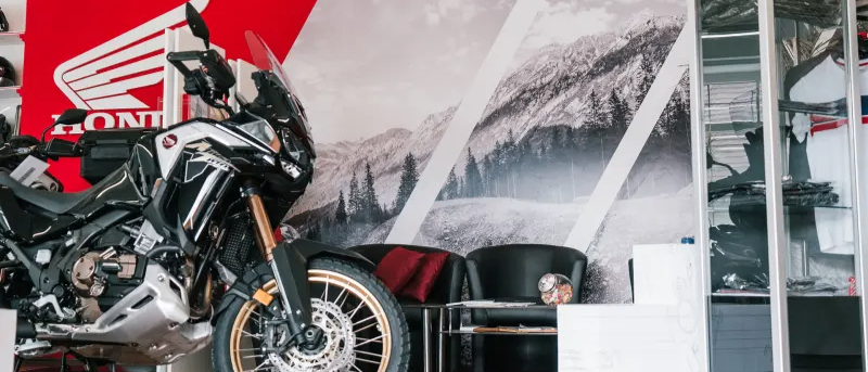 Ein Motorrad steht vor einer Wand mit einem Bergbild, zwei schwarzen Sesseln und einem Regal. Das Motorrad hat eine schwarz-silberne Farbe. Das Honda-Logo befindet sich an der Wand.
