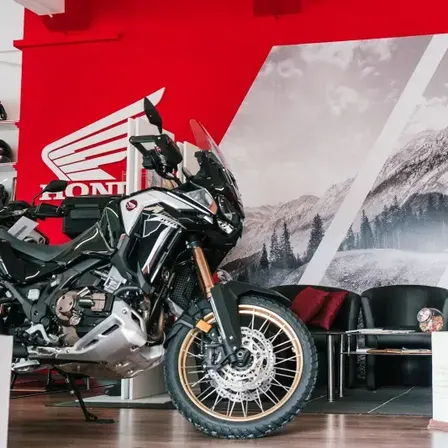 Ein schwarzes Honda-Motorrad steht in einem Ausstellungsraum mit einer roten Wand. Die Wand zeigt ein großes Wandbild einer Bergkette und ein großes Honda-Logo.
