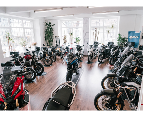 Ein Showroom voller verschiedener Motorräder, jedes auf einem Holzboden. Die Wände sind weiß, mit großen Fenstern und Topfpflanzen. Oben beleuchten Leuchtstofflampen den Raum.