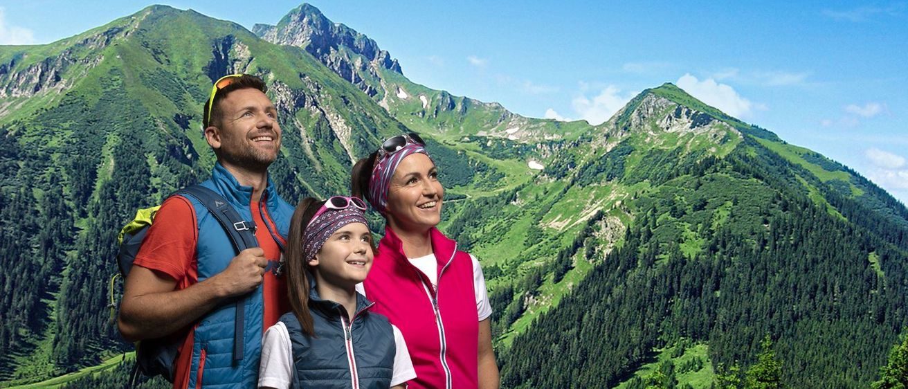 Eine dreiköpfige Familie steht vor einer Berglandschaft. Der Mann trägt einen Rucksack, die Frau hat eine Sonnenbrille auf dem Kopf und das Mädchen trägt ein Stirnband.