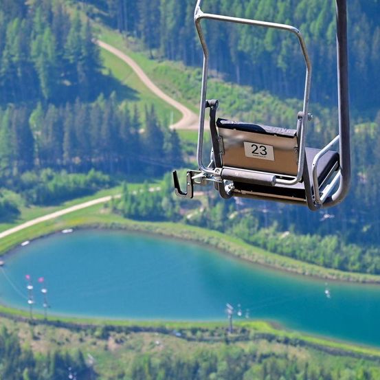 Ein Skiliftsitz mit der Nummer 23 schwebt über einem ruhigen See, umgeben von üppigen grünen Bäumen und einer gewundenen Straße.
