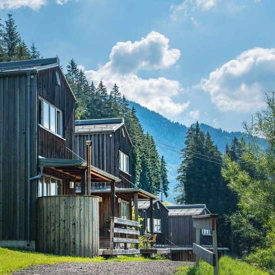 Ein Holzhaus mit Veranda und Fass steht auf einem grün bewachsenen Hügel, umgeben von hohen Bäumen und Bergen unter einem blauen Himmel mit Wolken.