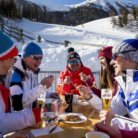 Eine Gruppe von Menschen in Winterkleidung genießt eine Mahlzeit an einem Tisch in einer verschneiten Outdoor-Umgebung. Sie tragen Wintermützen und Brillen, und es gibt Getränke und Essen auf dem Tisch. Im Hintergrund befindet sich ein verschneiter Berg.