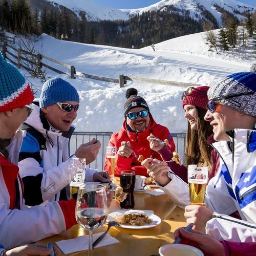 Eine Gruppe von Menschen in Winterkleidung genießt eine Mahlzeit an einem Tisch in einer verschneiten Outdoor-Umgebung. Sie tragen Wintermützen und Brillen, und es gibt Getränke und Essen auf dem Tisch. Im Hintergrund befindet sich ein verschneiter Berg.