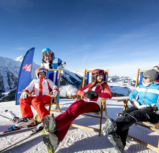 Eine vierköpfige Familie sitzt auf Stühlen auf einem verschneiten Berg, mit einer Person, die hinter ihnen steht. Sie tragen alle Winterkleidung und Skiausrüstung.