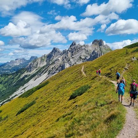 Eine Gruppe von Wanderern erklimmt einen grasbewachsenen Berghang unter einem blauen Himmel mit verstreuten Wolken. Der schmale Pfad führt zu einer felsigen Bergkette.
