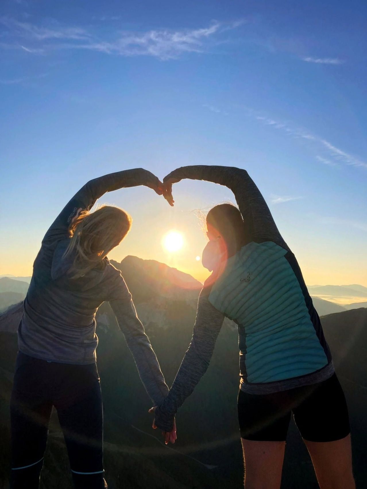 Two women form a heart shape with their arms on a mountain peak during sunset.