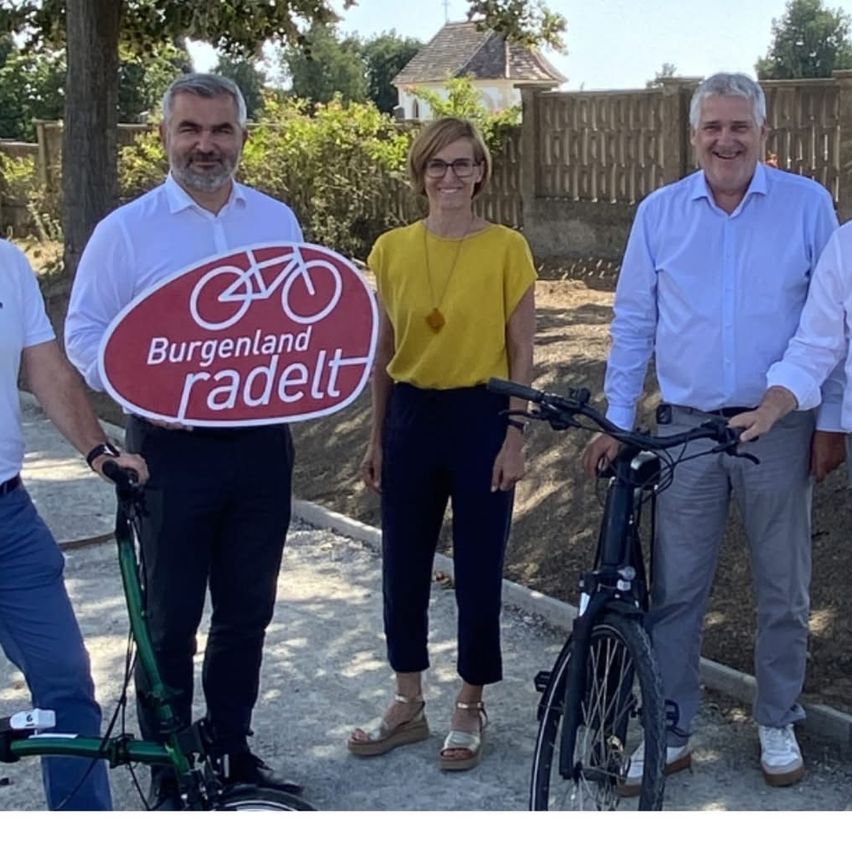 Vier Erwachsene posieren mit Fahrrädern für ein Foto im Freien. Ein Mann hält ein Schild mit der Aufschrift 'Burgenland radelt'. Die Gruppe lächelt.
