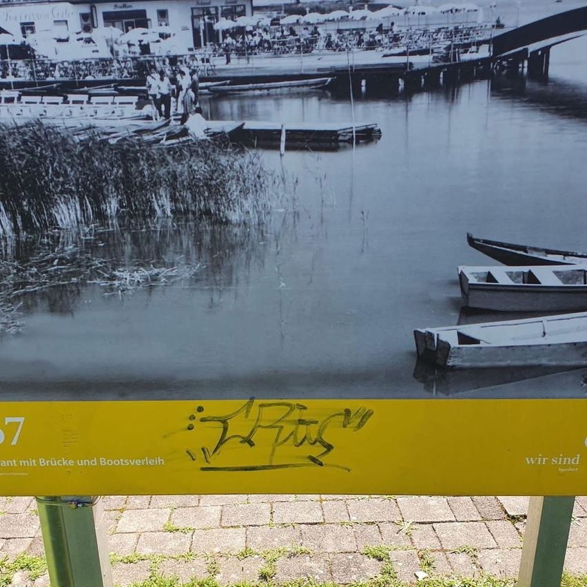 Ein gelbes Schild mit schwarzer Graffiti zeigt die Nummer '7'. Es ist auf einer Ziegeloberfläche platziert. Ein schwarz-weißes Bild zeigt Boote auf einem See und eine Brücke.