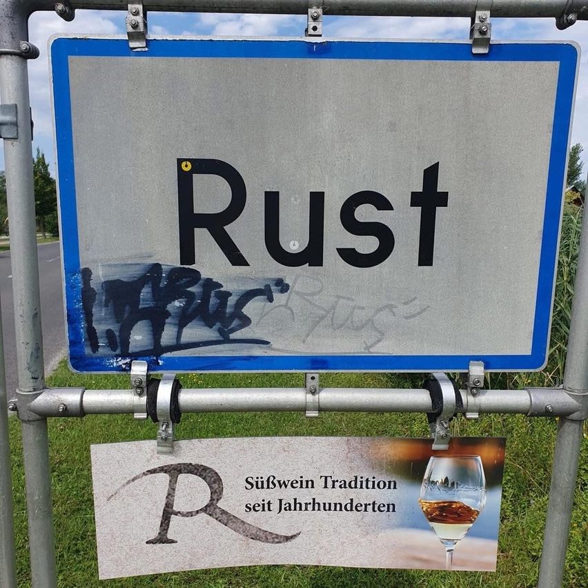 Ein Straßenschild mit der Aufschrift Rust und Graffiti. Darunter ist eine Werbung für Süßwein mit einem Bild eines Weinglases.