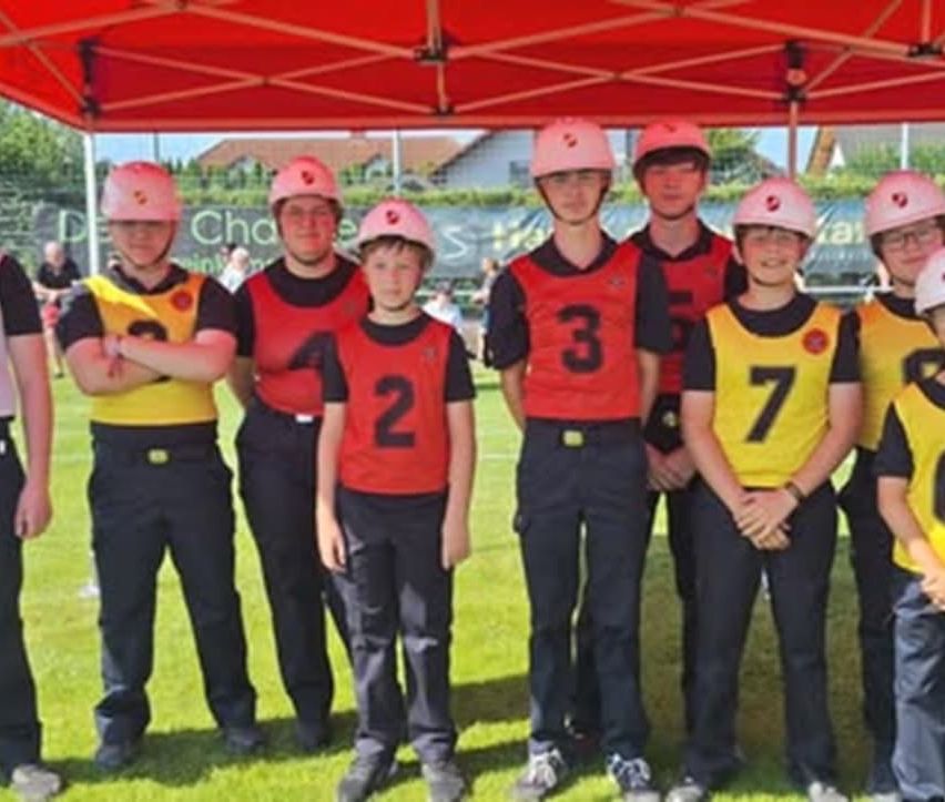 Eine Gruppe junger Personen in Helmen und nummerierten Trikots posiert für ein Foto auf einem Rasenfeld. Sie stehen unter einem roten Zelt.