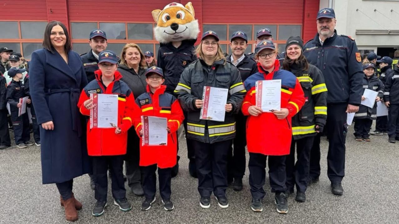 Eine Gruppe von Menschen, einschließlich Kindern, steht vor einer Feuerwache, trägt rote und blaue Uniformen. Sie halten Urkunden in den Händen, und ein Maskottchen ist ebenfalls anwesend.