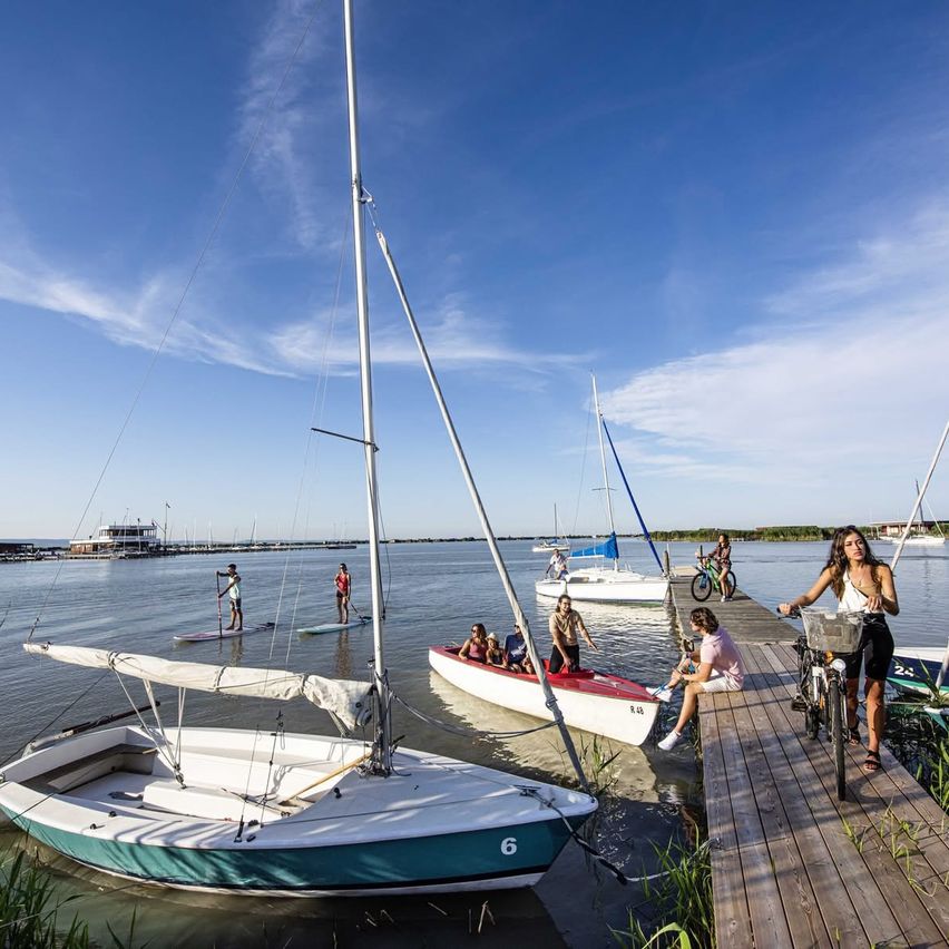 Eine Gruppe von Menschen genießt Wassersport an einem sonnigen Tag, mit Booten und Paddleboards im Wasser und anderen auf einem Holzsteg. Eine Frau fährt in der Nähe mit dem Fahrrad.